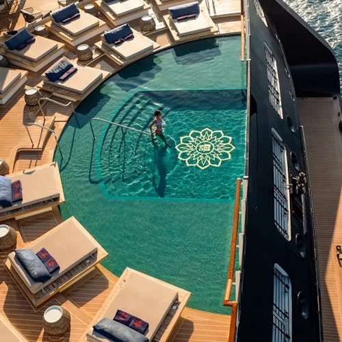 Luxury cruise ship pool deck with sun loungers and mosaic-tiled swimming pool overlooking the ocean.